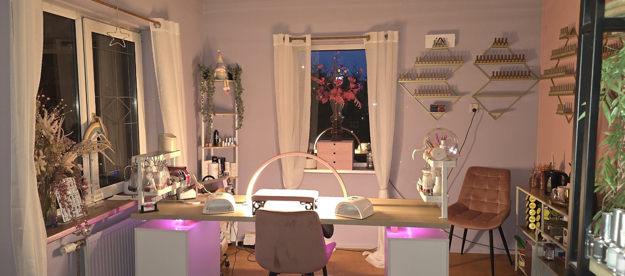 De studio van Pride Nails: de nageltafel met twee lampen, roze stoelen, gouden schapjes met lakjes aan de wand en een raam met bloemen